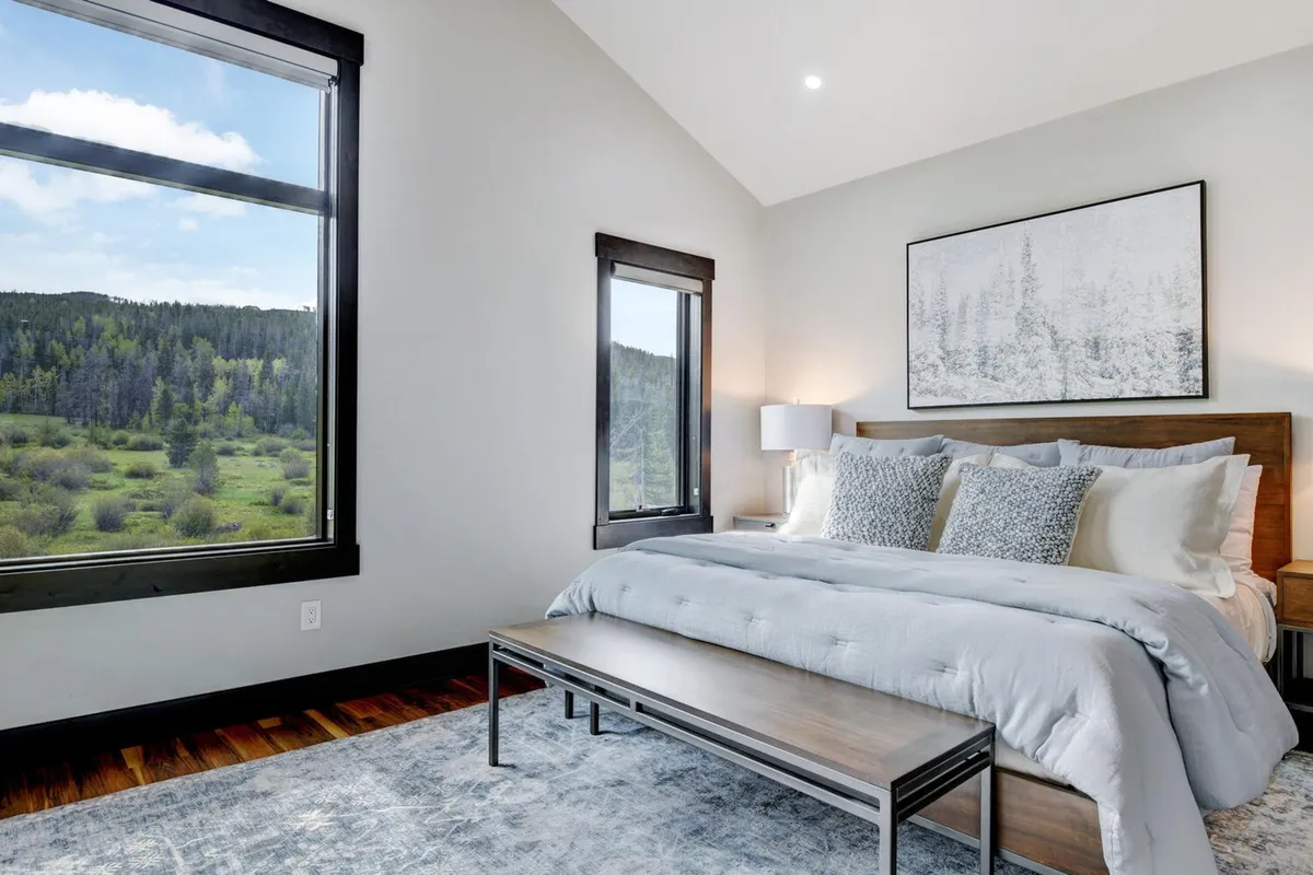 This image features a spacious, comfortable bedroom with a large window offering breathtaking mountain views. The bed is adorned with plush bedding, an art piece hangs above the headboard, and a wooden bench sits at the foot of the bed, creating a serene and inviting retreat.