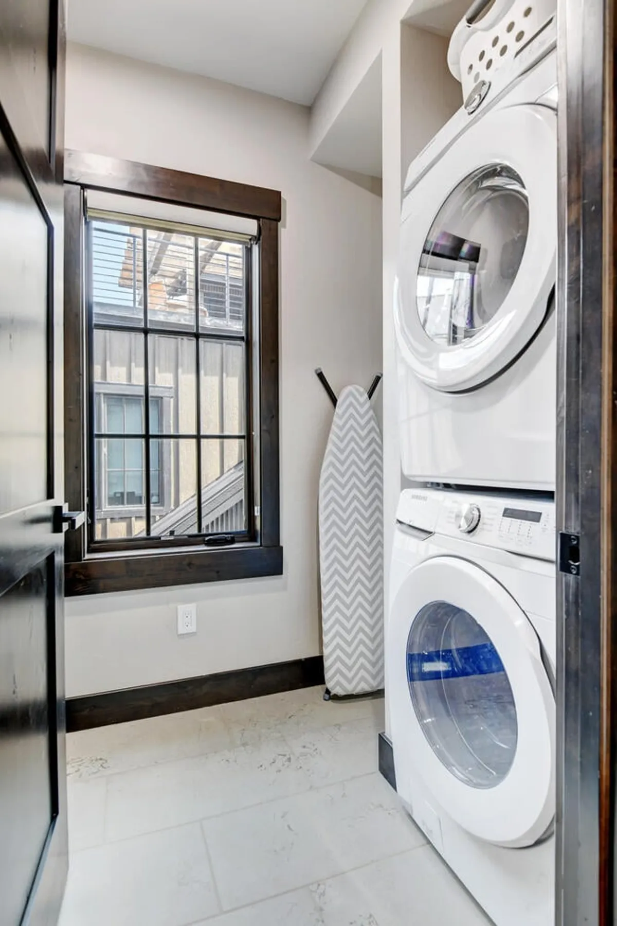 A tidy and practical laundry area with a stacked washer and dryer, a window letting in natural light, and an ironing board nearby. The space is organized, clean, and accessible.