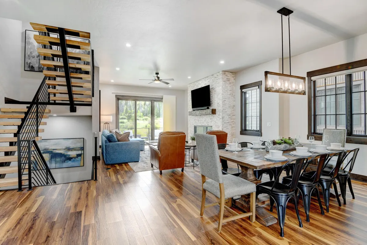 This image presents a welcoming, open living area with a staircase, a generous dining table, a spacious living zone, and large windows letting in abundant natural light. The arrangement suggests spaces for gathering, socializing, and relaxing, with the ambiance of a cozy mountain retreat.
