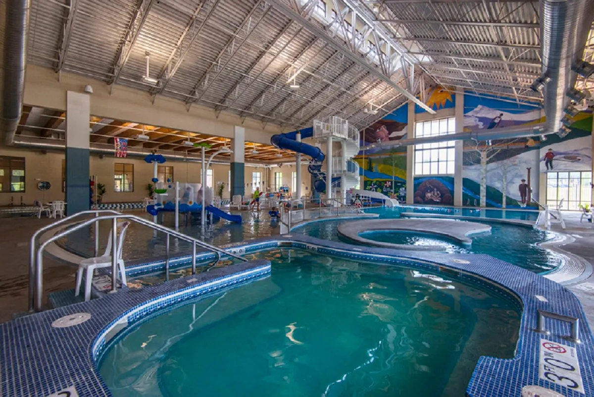 This interior image captures the lively atmosphere of a recreation center pool area, featuring a colorful mural, children playing, and a water slide. The space is filled with natural light, and the energetic scene emphasizes active family fun and community bonding.