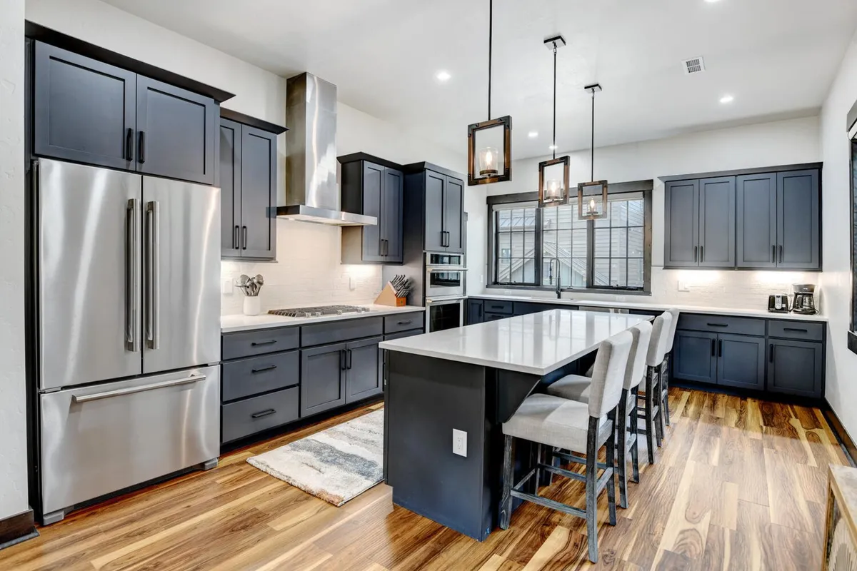 A modern kitchen with a prominent island bar seating four, surrounded by stainless steel appliances including a gas range stove and wine fridge. The space boasts dark cabinetry, expansive granite countertops, and large windows that flood the area with natural light, creating an inviting atmosphere for both cooking and socializing.