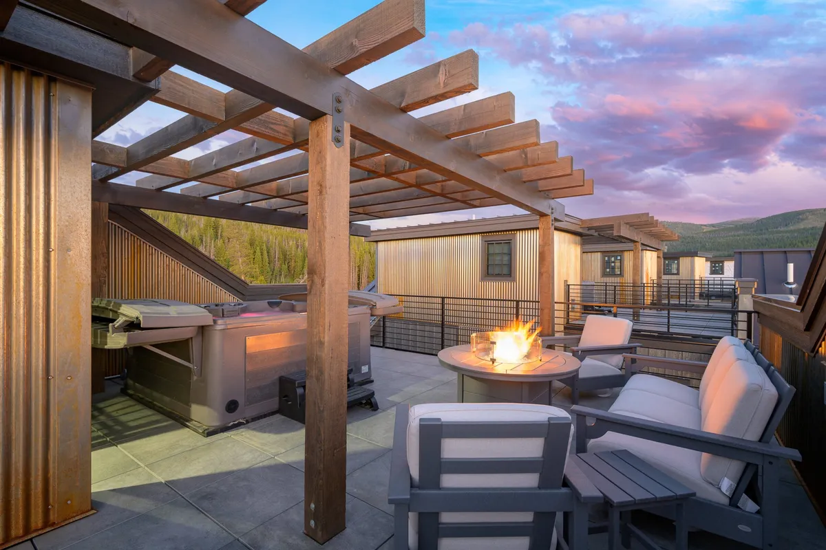 A spacious rooftop with a wooden pergola, hot tub, fire pit, and plush outdoor seating overlooks mountain vistas. The warm glow from the fire pit suggests a welcoming space for evening gatherings, while the hot tub promises relaxation.