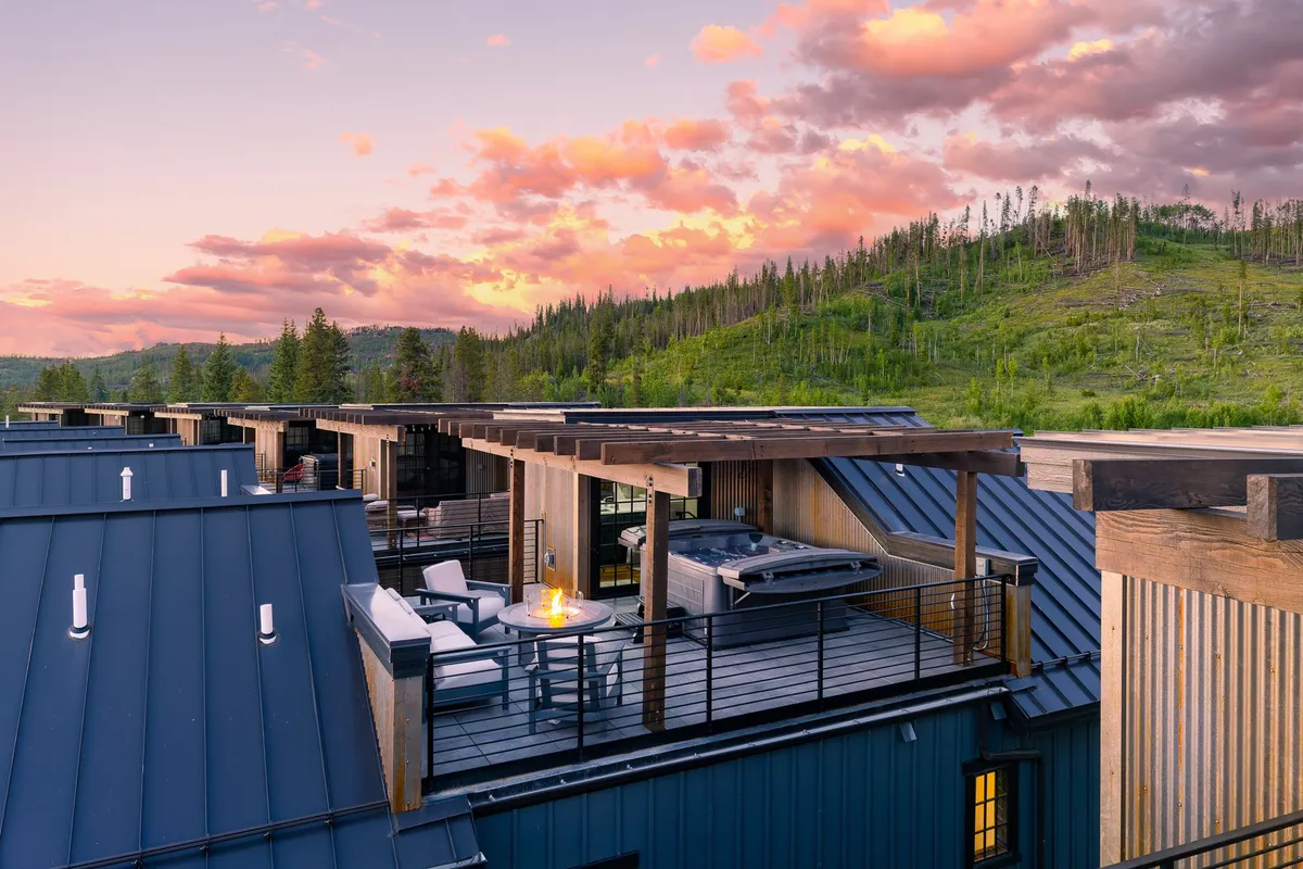 The hero image showcases a stunning rooftop deck overlooking majestic mountain vistas, featuring a modern hot tub, cozy fire pit, and comfortable lounge seating. The setting is perfect for unwinding under twinkling stars, enjoying sunset views, and gathering with friends or family in a serene alpine atmosphere.