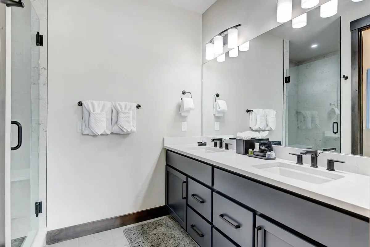 A modern, tranquil bathroom with double vanities, a large mirror, elegant light fixtures, and a spacious shower. The mood is calm and sophisticated, ideal for relaxation and privacy.