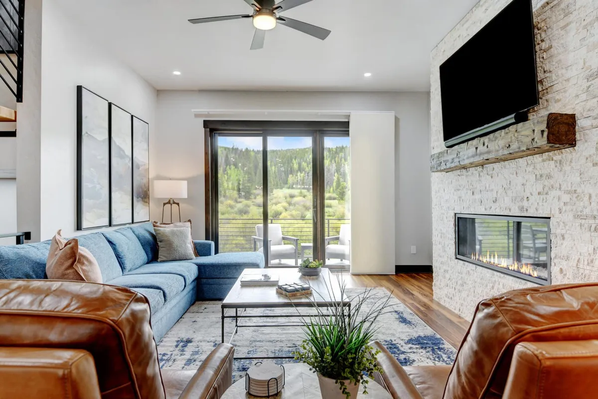 A stylish living room featuring a blue sofa facing a doublewide fireplace, large windows framing beautiful views, a coffee table, and decorative plant accents. The setting feels plush, warm, and ideal for gathering after outdoor adventures.