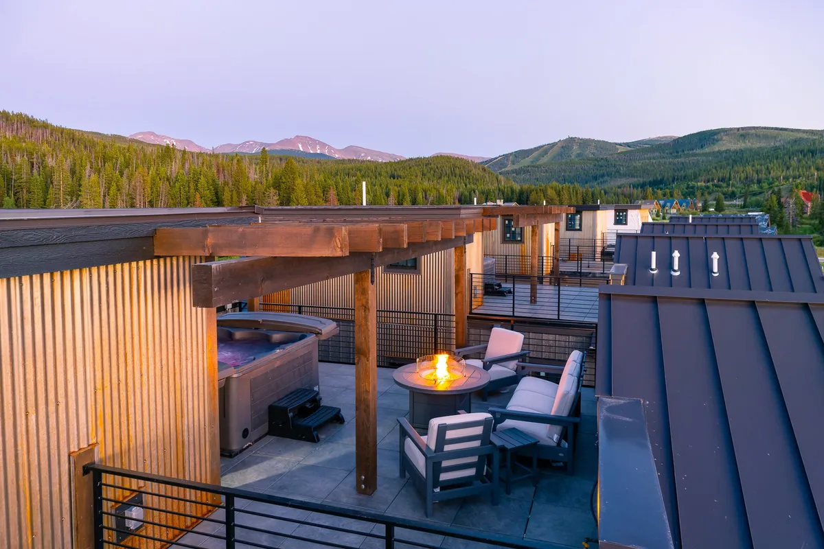 This image features a luxurious rooftop patio equipped with a private hot tub, a modern fire pit, a comfortable outdoor seating area, and sweeping views of the mountain landscape at sunset. The space is styled for both relaxation and entertaining.