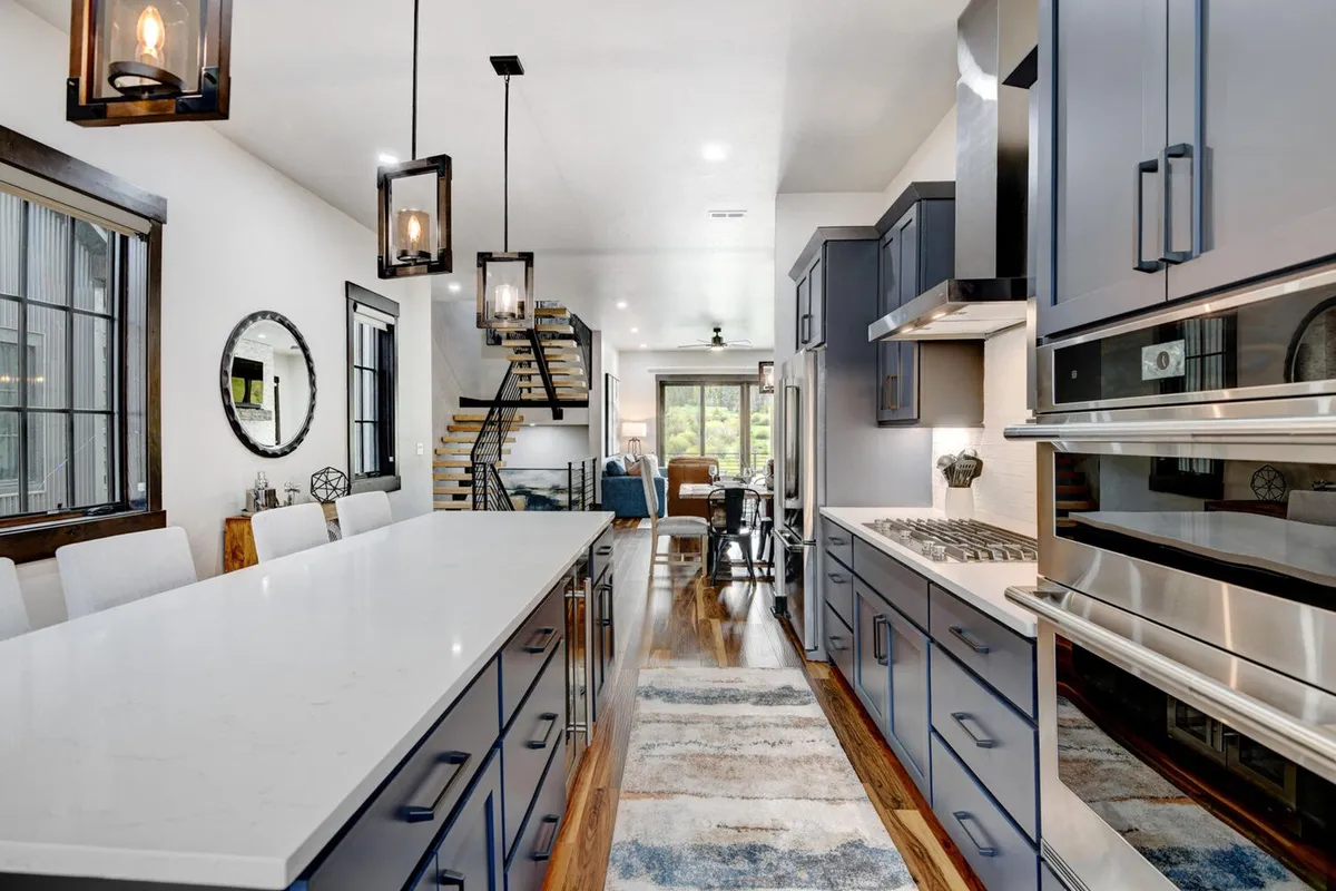 An expansive, welcoming kitchen highlighted by a large kitchen island, shiny stainless steel appliances, and modern lighting fixtures. The backdrop features unique touches such as a spiral staircase and decorative mirror, enhancing the kitchen’s luxurious and functional appeal for cooking and entertaining.